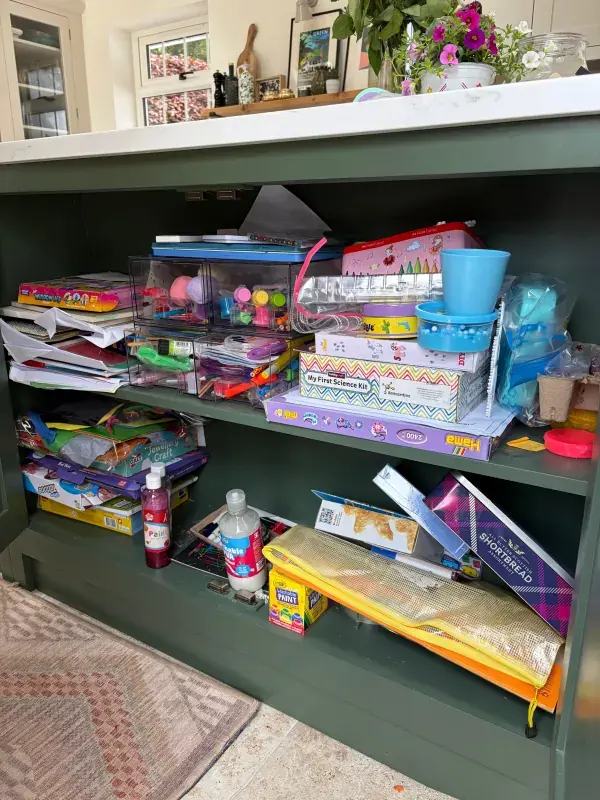 Children's craft cupboard before organising, with art kits, paper and paints piled on the shelves